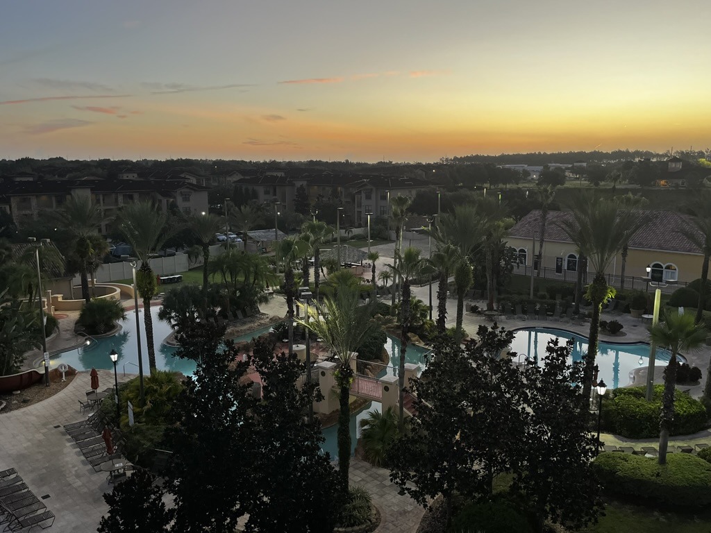 A serene outdoor pool area surrounded by palm trees at sunset, with residential buildings in the background and a calm, spacious atmosphere.
