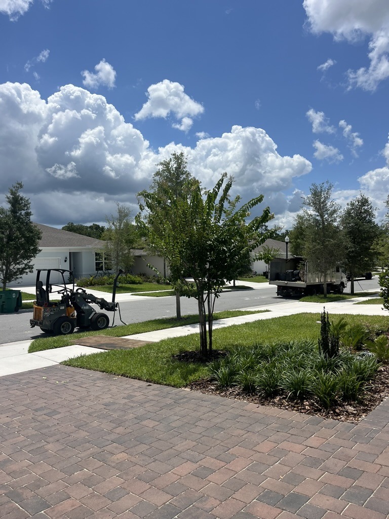 A sunny suburban street with a landscaping vehicle on the road, surrounded by green lawns and trees under a blue, cloudy sky.