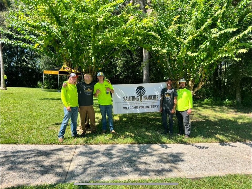 Five people in safety gear stand by a "Saluting Branches" volunteer banner, surrounded by green trees, on a sunny day.