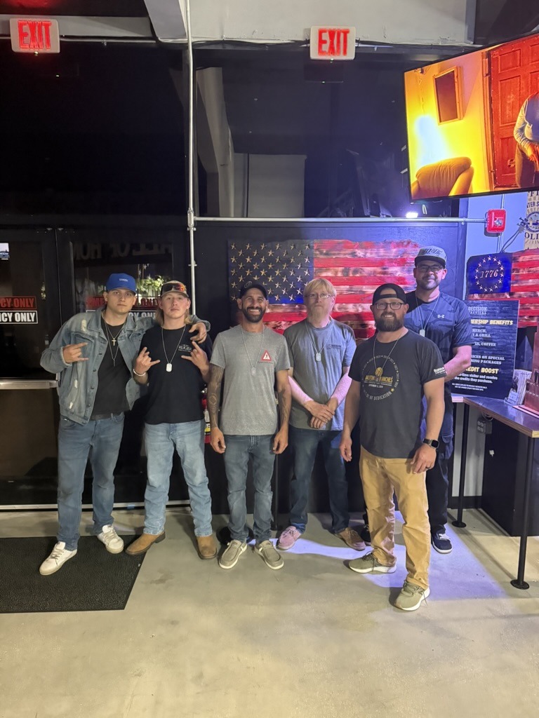 Six people posing indoors by a stylized American flag artwork. Bright lighting, exit signs visible. Casual attire suggests a relaxed gathering.
